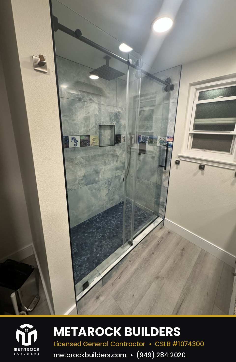 Shower remodel in Orange County by Metarock Builders, completed after photo with blue-gray onyx-look tile, pebble mosaic floor, matte black hardware, and sliding glass door
